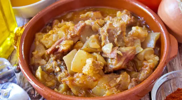 Guiso de cap i pota en cazuela de barro, con patatas y trozos de carne, rodeado de ingredientes como aceite, pimentón y tomates.