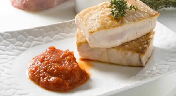 Dos trozos de atún a la plancha, dorados y jugosos, apilados sobre un plato blanco con una porción de salsa de tomate y una ramita de tomillo.