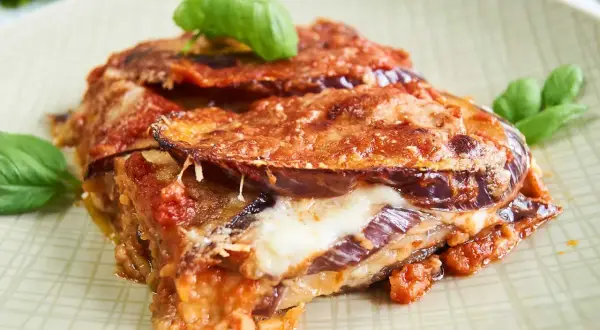Un trozo de parmigiana di melanzane, con capas de berenjena, queso fundido y salsa de tomate, adornado con hojas de albahaca fresca.