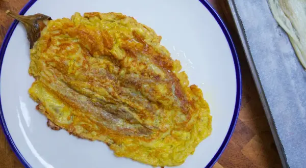 Tortilla de berenjena dorada y jugosa, servida en plato blanco con borde azul. Al lado, una bandeja con papel de horno y más berenjena.