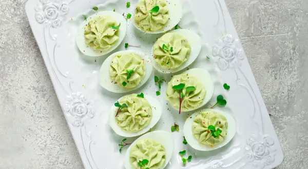 Deliciosos huevos rellenos de aguacate, decorados con brotes verdes y especias, servidos en una bandeja blanca con relieve floral.
