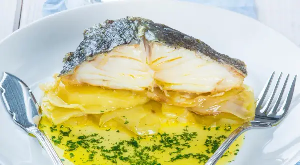 Merluza al horno con patatas, jugosa y dorada, servida sobre una cama de patatas y cebolla caramelizada, bañada en aceite de oliva y perejil picado.