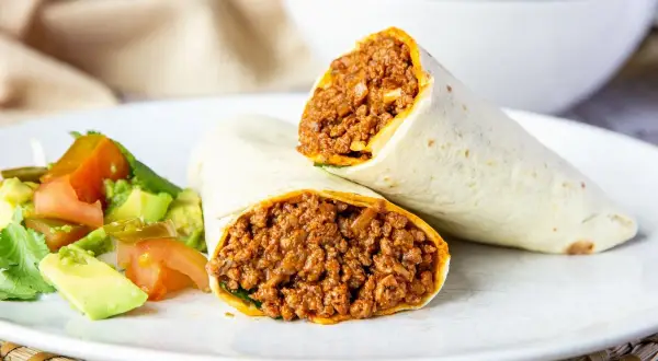 Dos burritos de carne picada, cortados por la mitad, servidos con una ensalada fresca de aguacate y tomate.