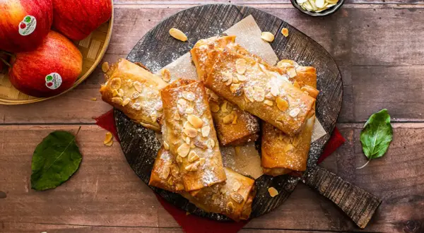 Deliciosos rollos de strudel de manzana dorados, espolvoreados con azúcar glas y almendras fileteadas, listos para disfrutar.