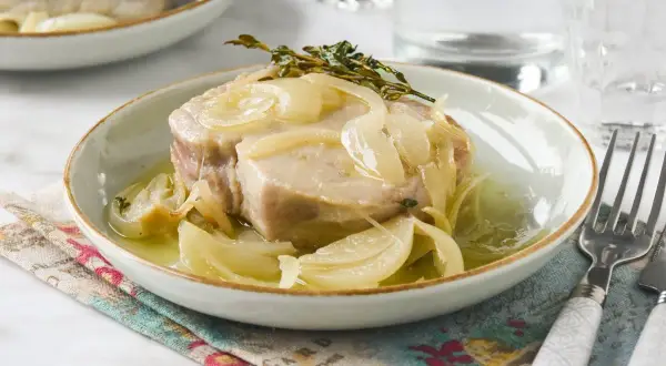 Un trozo de atún en escabeche con cebolla y tomillo, servido en un plato blanco con un tenedor al lado.