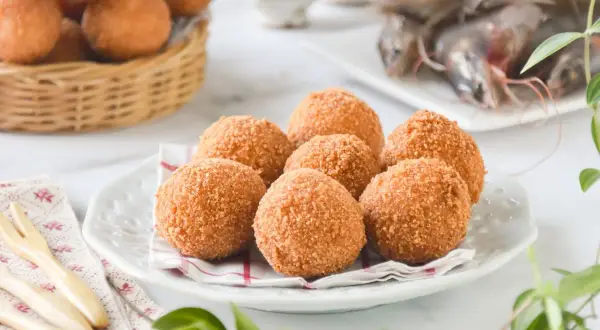Croquetas de gambas doradas y crujientes en un plato blanco, con más en una cesta de mimbre al fondo y gambas frescas a la derecha.