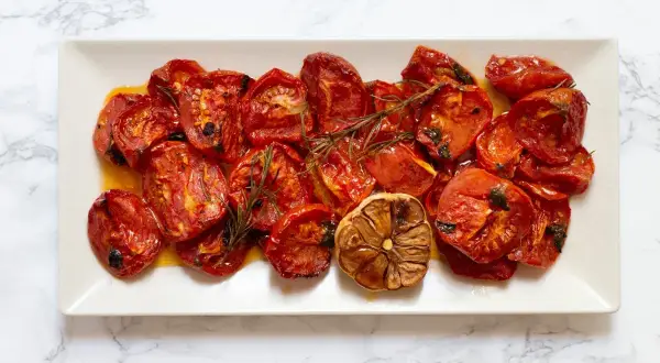 Tomates al horno, jugosos y caramelizados, con ajo asado y romero, servidos en una bandeja blanca sobre mármol.