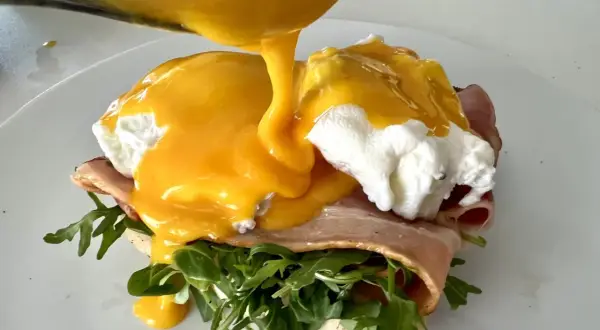 Un delicioso uovo alla benedict, con huevo escalfado, jamón, rúcula y salsa holandesa cremosa vertiéndose sobre él.