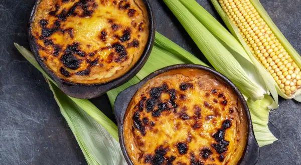 Dos cazuelas de pastel de choclo recién horneado, con su dorada y caramelizada cubierta, junto a mazorcas de maíz fresco.