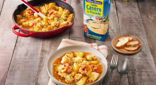 Delicioso rape a la marinera con patatas y gambas, servido en plato y sartén, junto a caldo de pescado y pan.