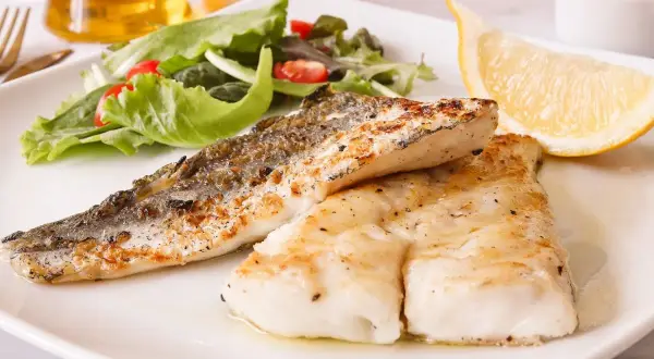 Deliciosa merluza a la plancha con piel crujiente, acompañada de ensalada fresca y un gajo de limón.