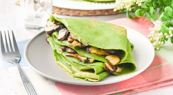 Deliciosos crepes de espinacas rellenos de champiñones salteados, servidos en un plato gris con un tenedor al lado.