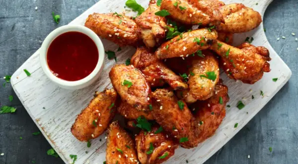 Alitas de pollo al horno crujientes, doradas y espolvoreadas con sésamo y perejil, servidas con salsa roja en un cuenco.