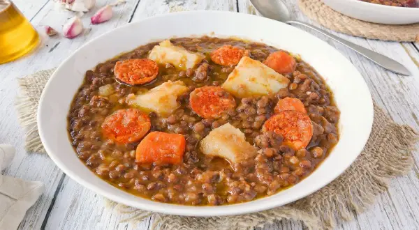 Un plato humeante de lentejas con chorizo, patatas y zanahoria, servido en un cuenco blanco sobre una mesa de madera.