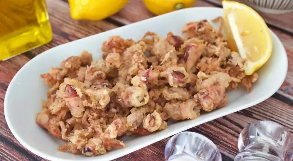 Crujientes puntillas de calamar fritas en un plato blanco, acompañadas de limón, aceite y saleros.