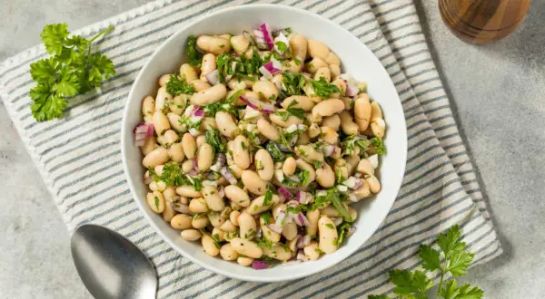 Deliciosa ensalada de judías blancas con cebolla morada y perejil fresco, servida en un cuenco blanco sobre un mantel a rayas.