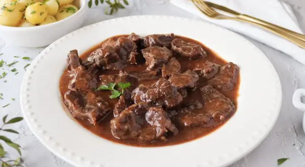 Delicioso morcillo de ternera estofado en salsa, servido en plato blanco con patatas cocidas y perejil picado.