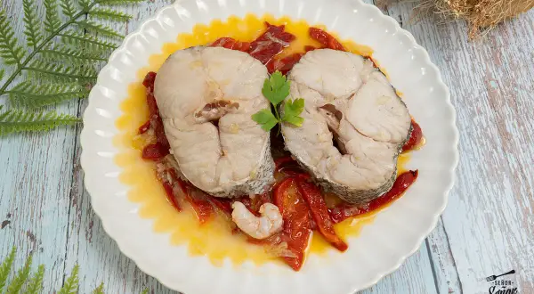 Dos rodajas de merluza a la riojana, servidas sobre pimientos rojos y una gamba, decoradas con perejil.