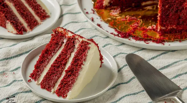 Porción de pastel red velvet con capas de bizcocho rojo y crema blanca. Otro trozo y el pastel entero en el fondo.
