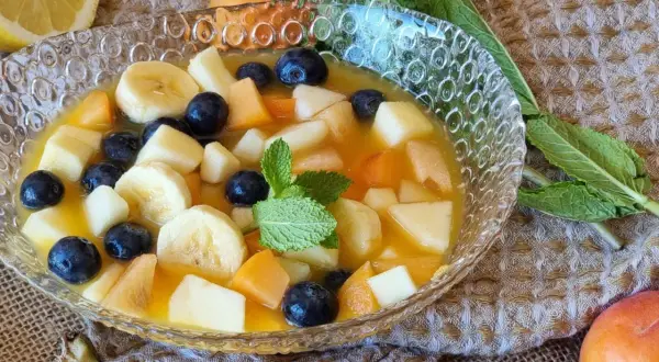 Deliciosa macedonia de frutas con plátano, arándanos, melocotón y manzana en un cuenco de cristal, decorada con menta.