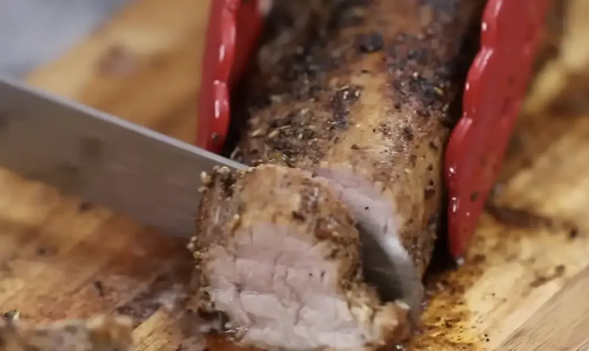 Un jugoso solomillo de cerdo al horno, recién cortado en una tabla de madera. Las pinzas rojas sujetan la carne mientras el cuchillo la lonchea.