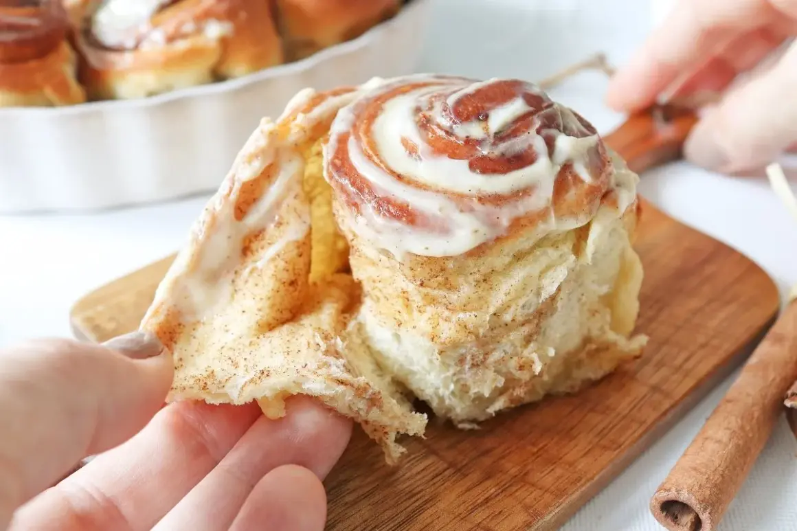 Un rollo de canela recién horneado, cubierto con glaseado cremoso, se desmenuza suavemente al ser tomado.