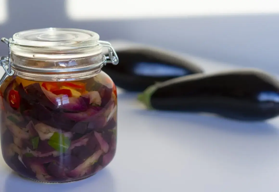 Tarro de berenjenas en escabeche con trozos de pimiento rojo y hierbas. Dos berenjenas frescas en el fondo.