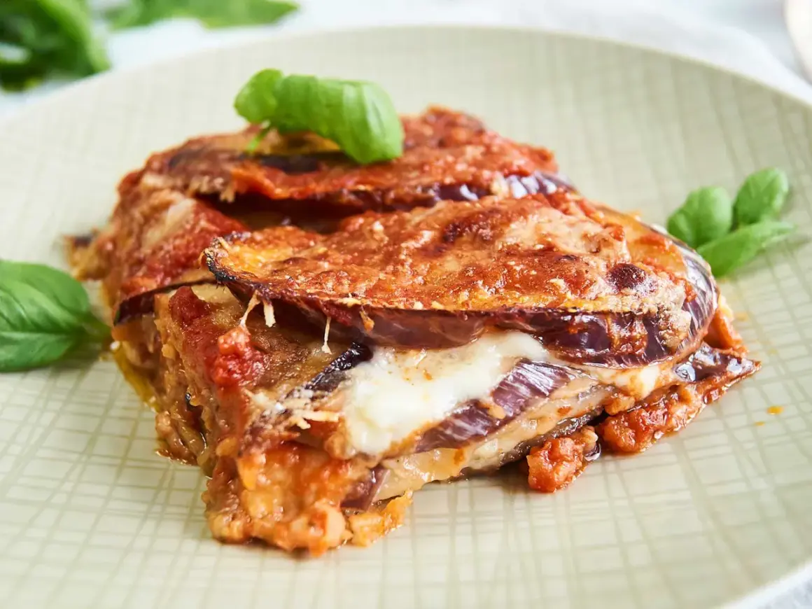 Un trozo de parmigiana di melanzane, con capas de berenjena, queso fundido y salsa de tomate, adornado con hojas de albahaca fresca.
