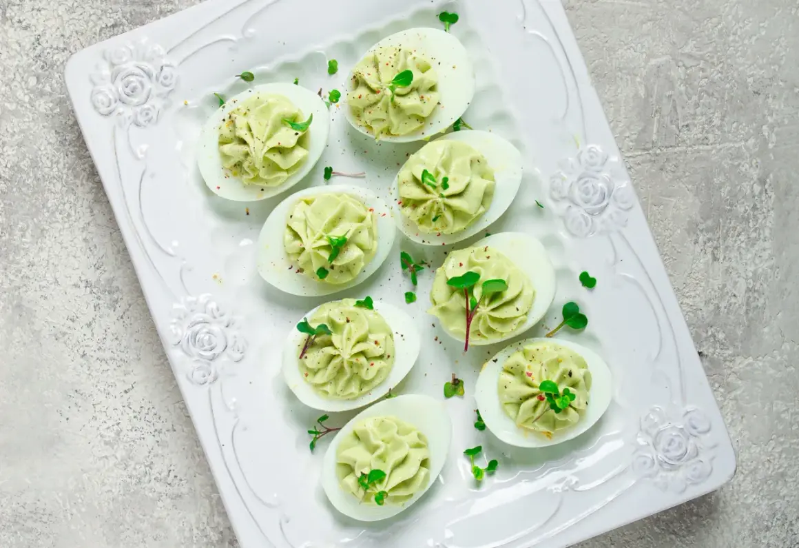 Deliciosos huevos rellenos de aguacate, decorados con brotes verdes y especias, servidos en una bandeja blanca con relieve floral.