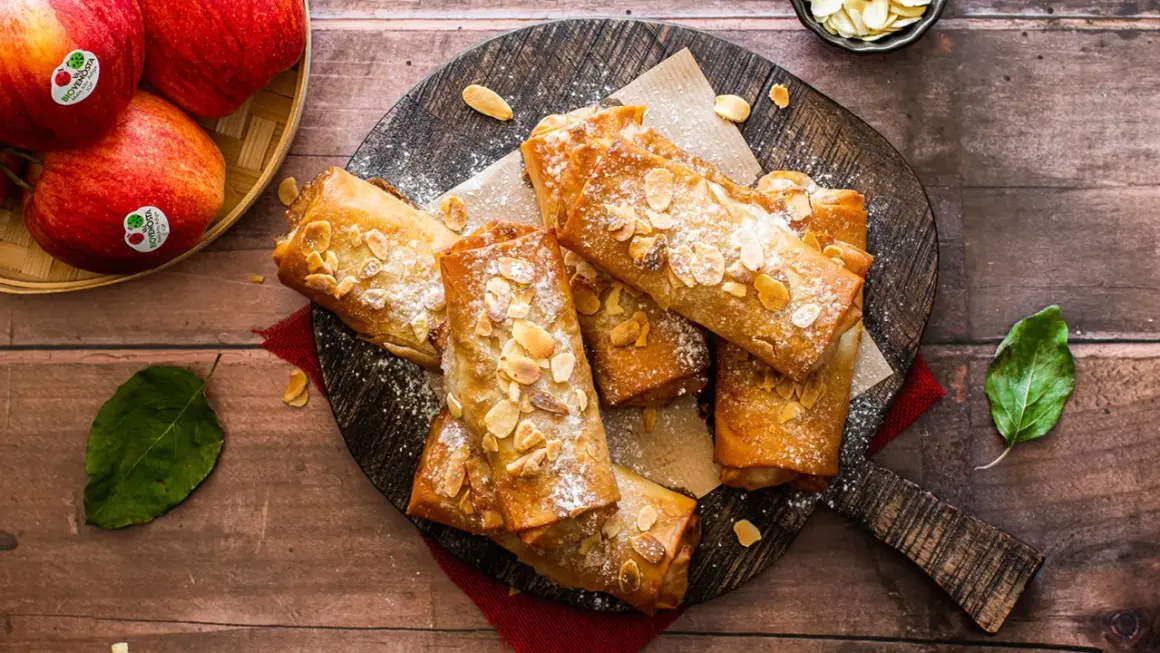 Deliciosos rollos de strudel de manzana dorados, espolvoreados con azúcar glas y almendras fileteadas, listos para disfrutar.