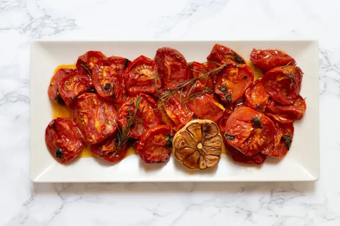 Tomates al horno, jugosos y caramelizados, con ajo asado y romero, servidos en una bandeja blanca sobre mármol.