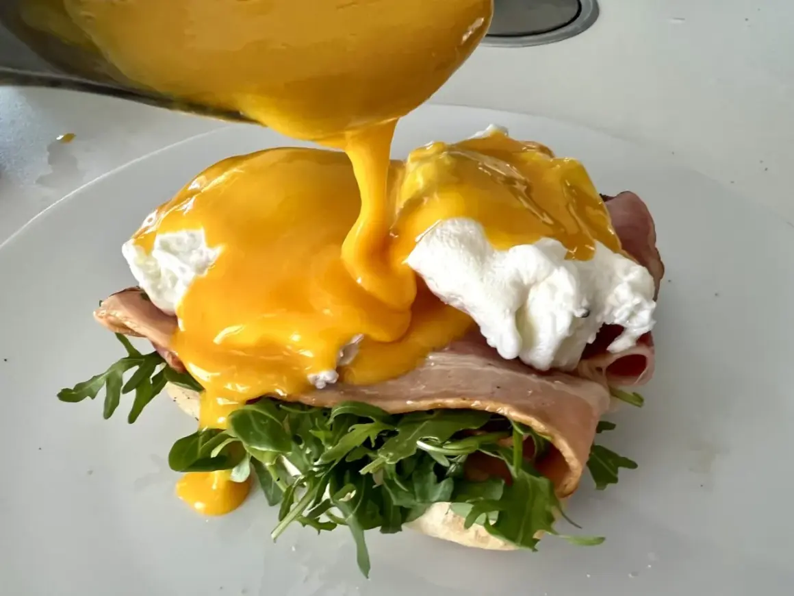 Un delicioso uovo alla benedict, con huevo escalfado, jamón, rúcula y salsa holandesa cremosa vertiéndose sobre él.