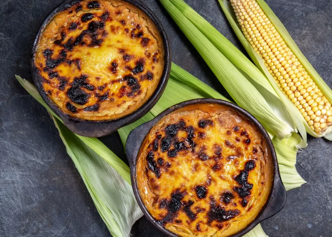 Dos cazuelas de pastel de choclo recién horneado, con su dorada y caramelizada cubierta, junto a mazorcas de maíz fresco.