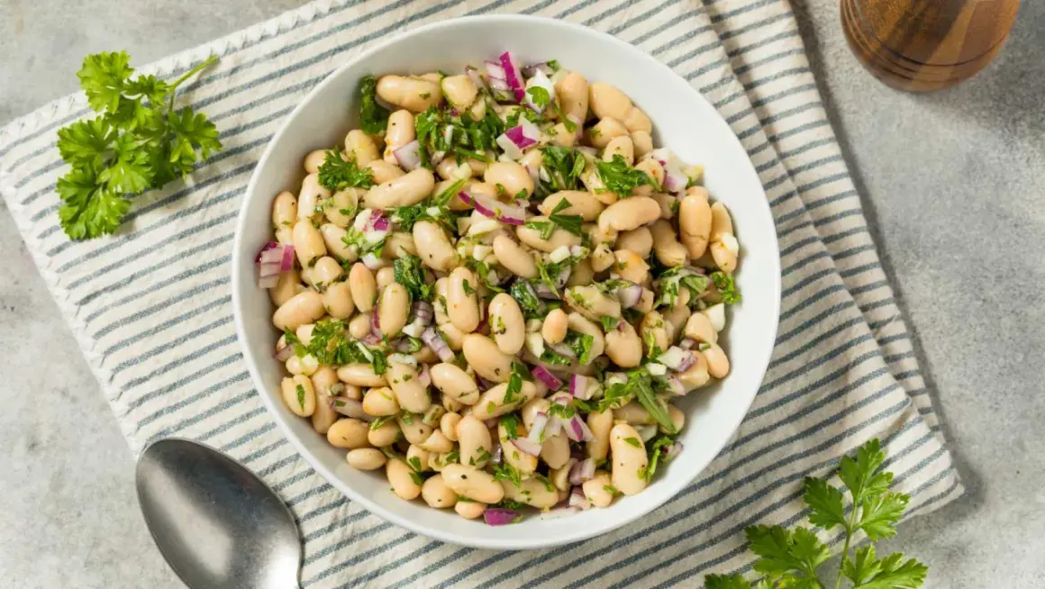 Deliciosa ensalada de judías blancas con cebolla morada y perejil fresco, servida en un cuenco blanco sobre un mantel a rayas.