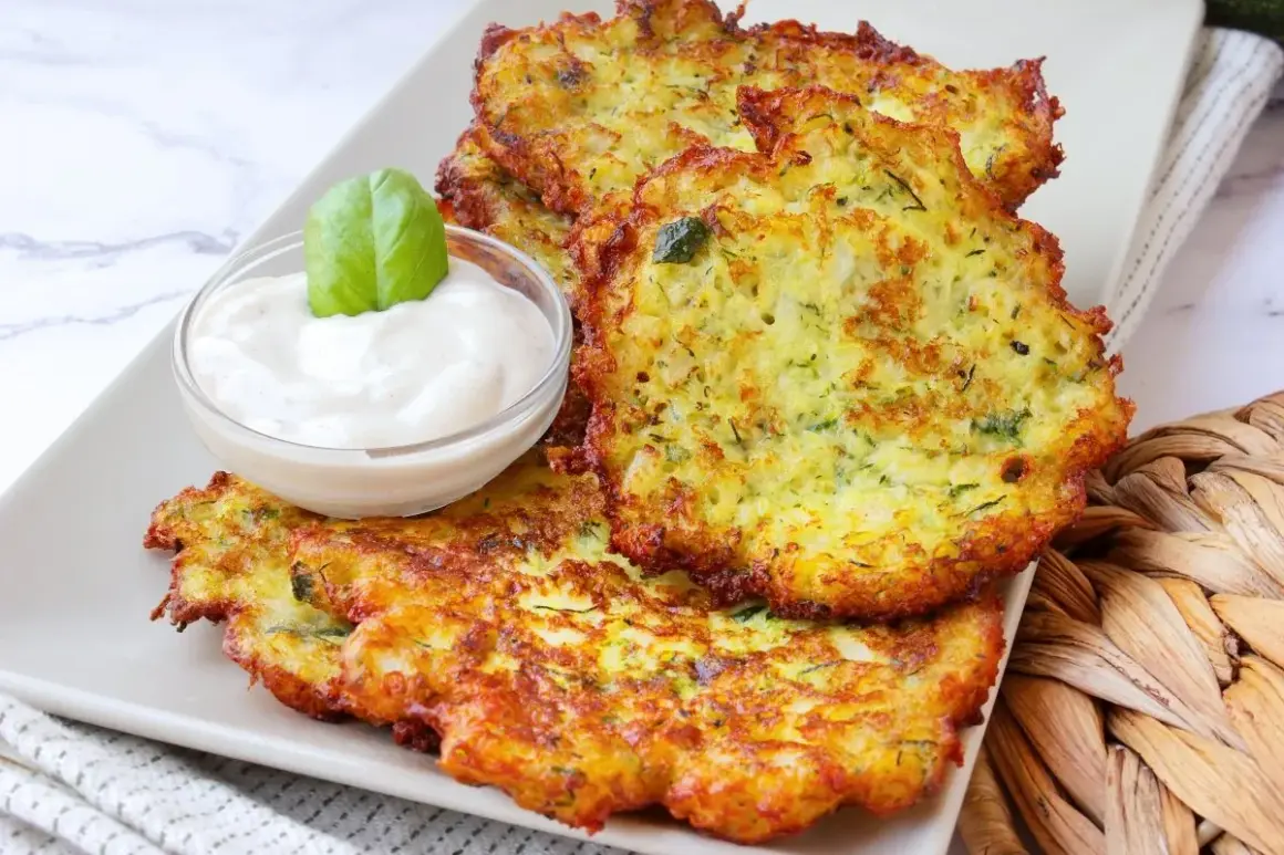 Tortitas de calabacín doradas y crujientes, servidas con una cremosa salsa y decoradas con una hoja de albahaca.