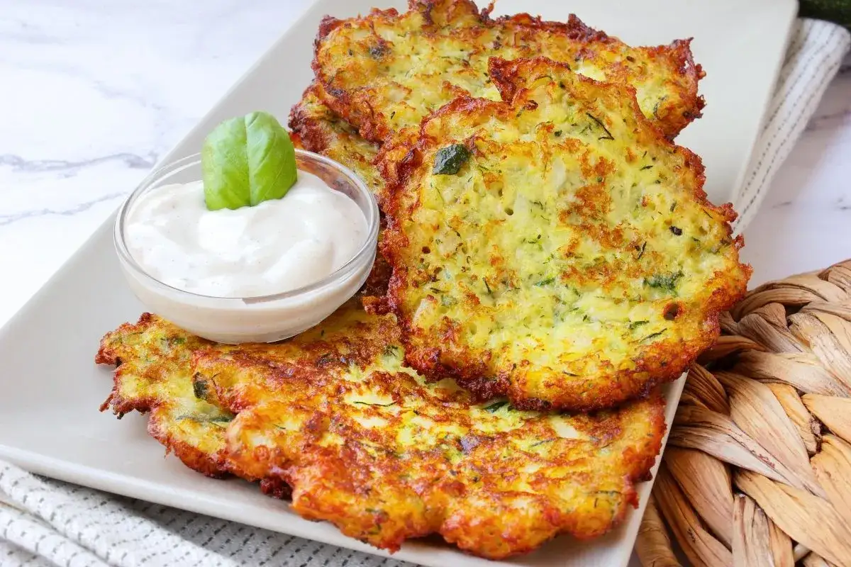 Tortitas de calabac&iacute;n doradas y crujientes, servidas con una cremosa salsa y decoradas con una hoja de albahaca.