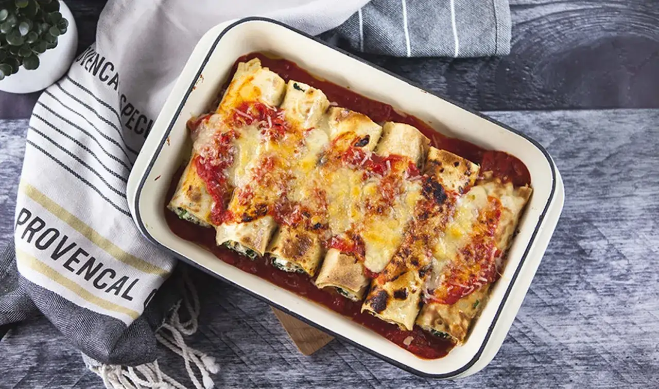 Rollo de crepes de espinacas gratinados con queso y salsa de tomate en una bandeja.