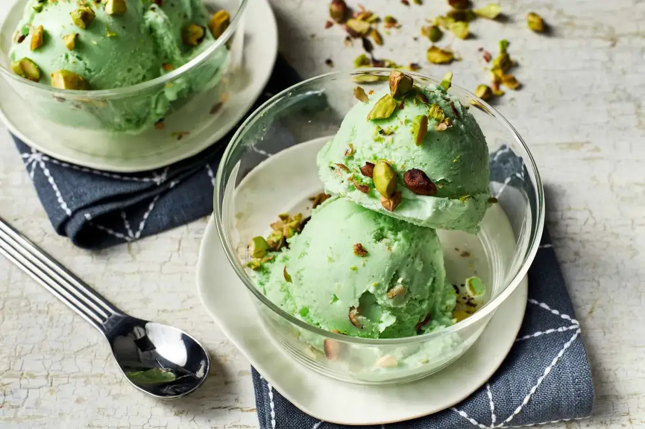 Dos bolas de cremoso helado de pistacho, adornadas con trozos de pistacho, en un cuenco de cristal.