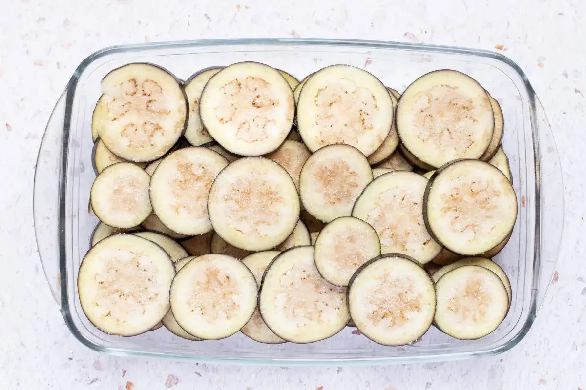 Rodajas de berenjenas listas para preparar berenjenas en escabeche, cubiertas con sal en un recipiente de vidrio.