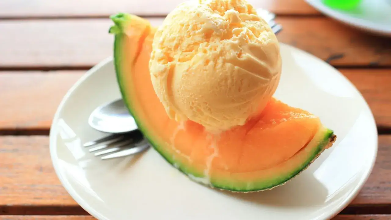Una bola de helado de melón se derrite sobre una rebanada de melón fresco en un plato blanco.