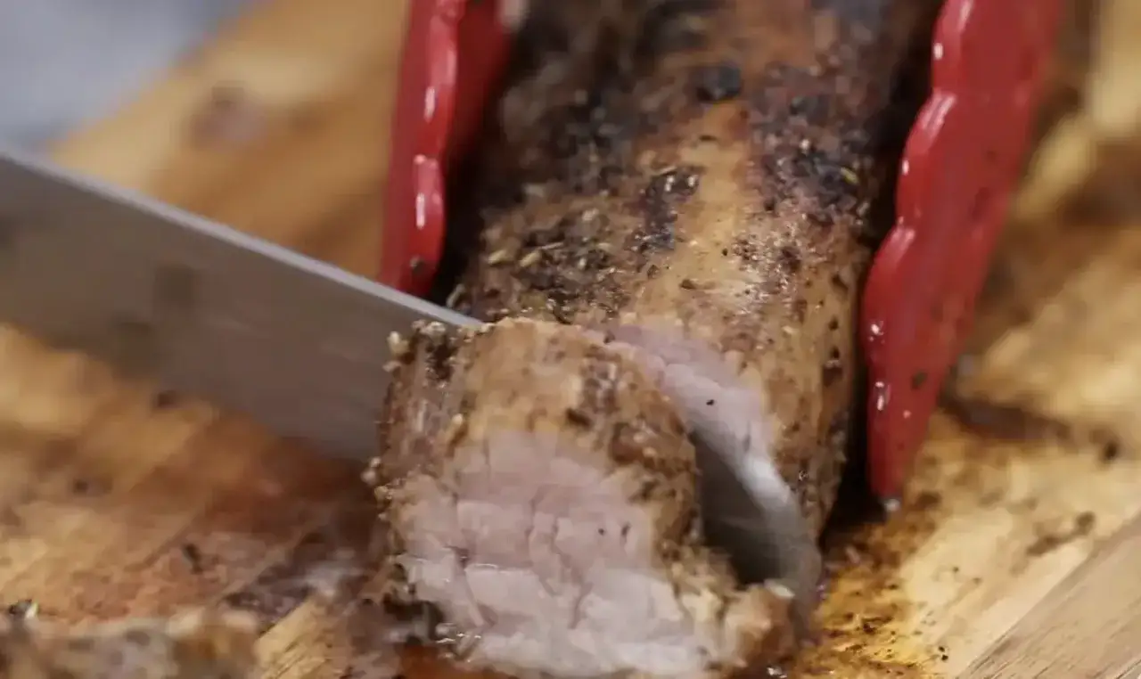 Un jugoso solomillo de cerdo al horno, recién cortado en una tabla de madera. Las pinzas rojas sujetan la carne mientras el cuchillo la rebana.