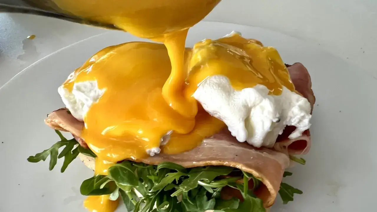 Un delicioso uovo alla benedict con huevo escalfado, tocino y r&uacute;cula, ba&ntilde;ado en salsa holandesa cremosa.