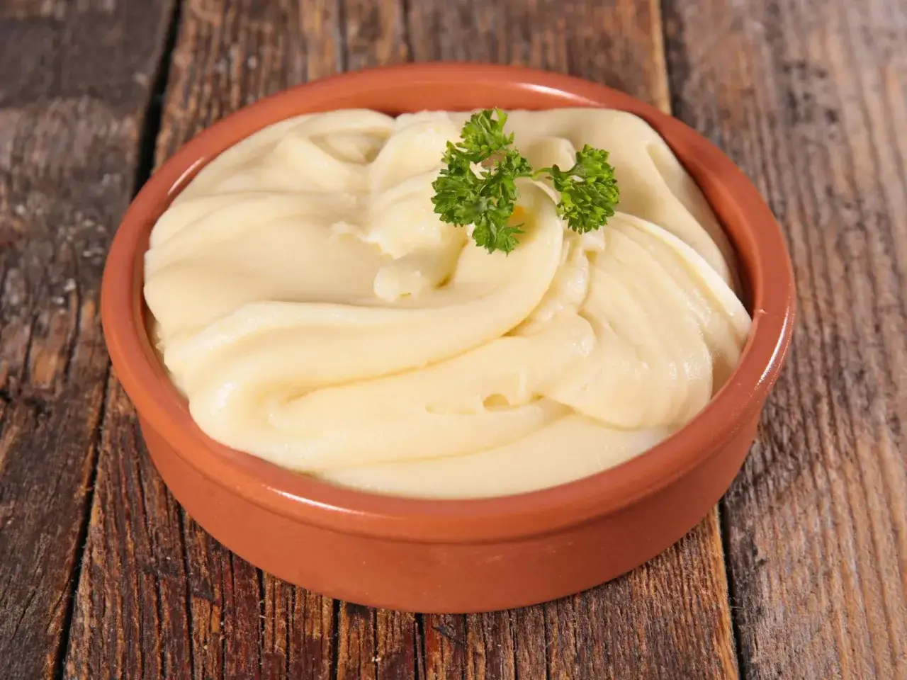 Un cuenco de barro con cremoso pure de patatas, adornado con una ramita de perejil fresco, sobre una mesa de madera rústica.