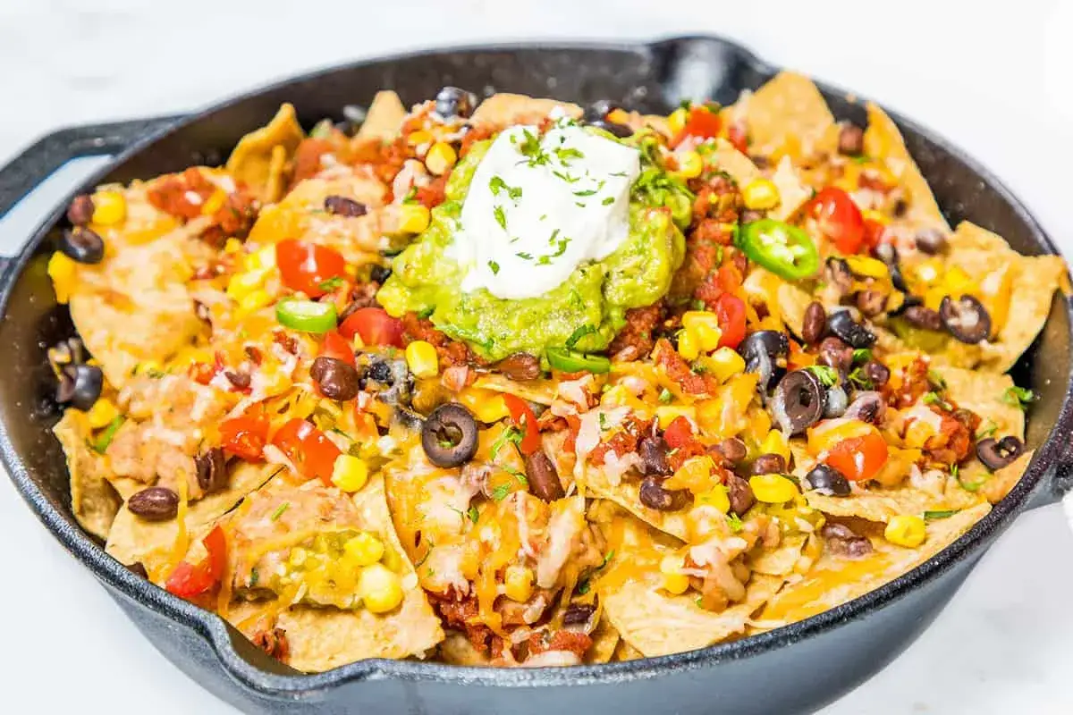 Nachos con queso rebosantes en sartén de hierro fundido, con guacamole, frijoles, maíz y jalapeños.