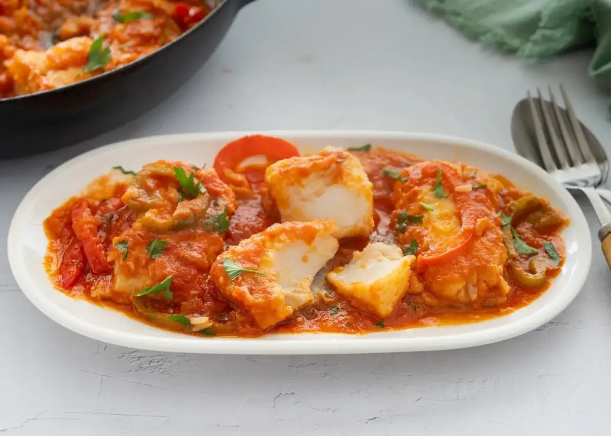 Bacalao con tomate, pimientos y perejil, servido en plato blanco. Un plato casero y delicioso.