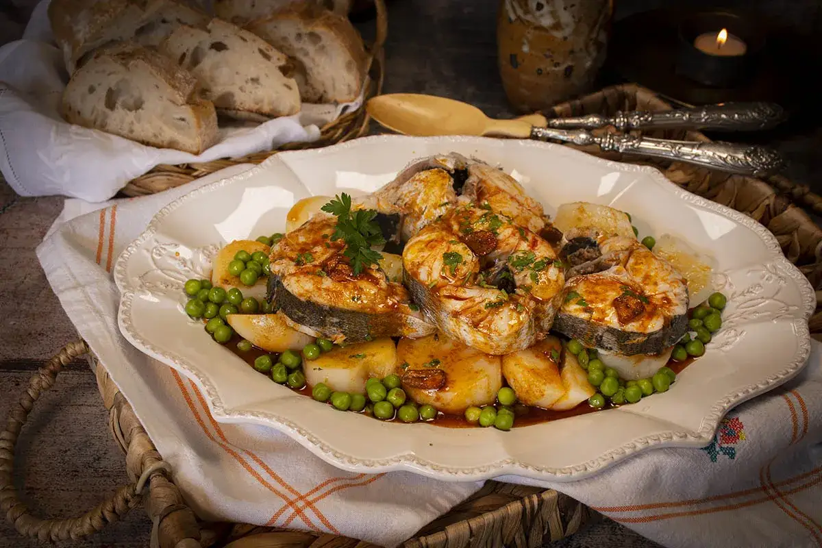 Merluza a la gallega servida con patatas y guisantes, decorada con perejil fresco. Pan r&uacute;stico y una vela completan la escena.