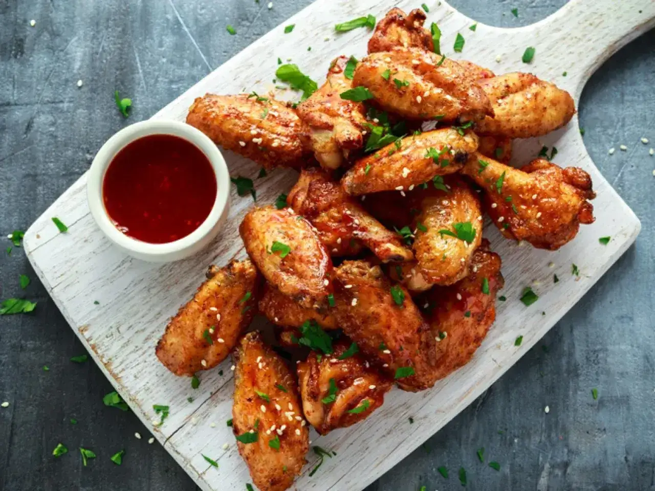 Alitas de pollo al horno crujientes, doradas y espolvoreadas con sésamo y perejil, acompañadas de una salsa roja.