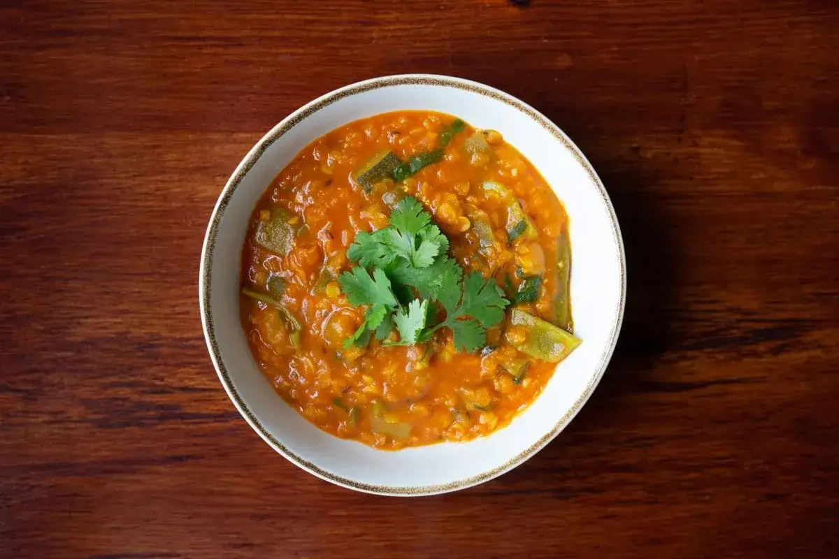 Un reconfortante plato de lentejas al curry, con cilantro fresco, servido en un cuenco blanco sobre una mesa de madera oscura.