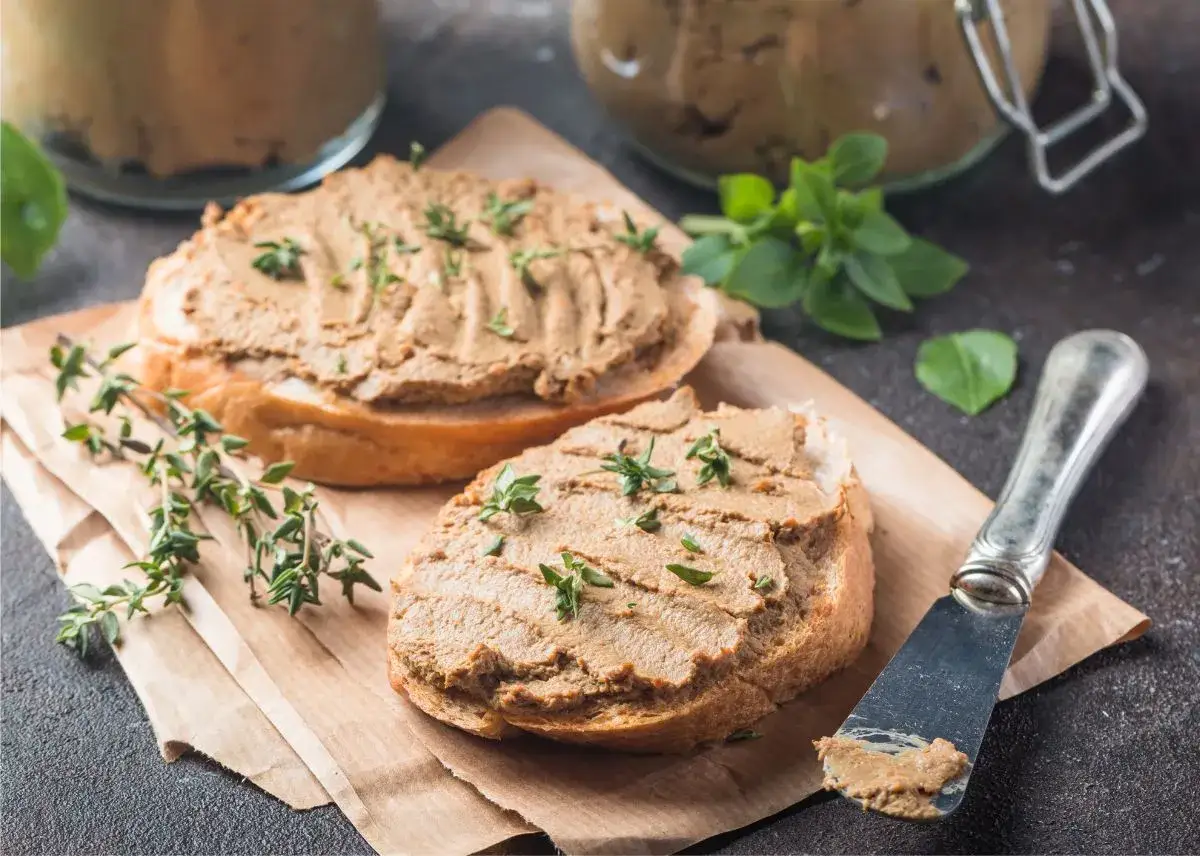 Dos rebanadas de pan con paté de hígado de pollo, decoradas con tomillo fresco. Un cuchillo con paté y hierbas verdes al fondo.