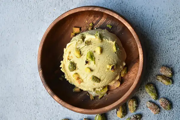 Cremoso helado de pistacho servido en un cuenco de madera, adornado con trozos de pistacho y acompañado de más pistachos enteros esparcidos alrededor.